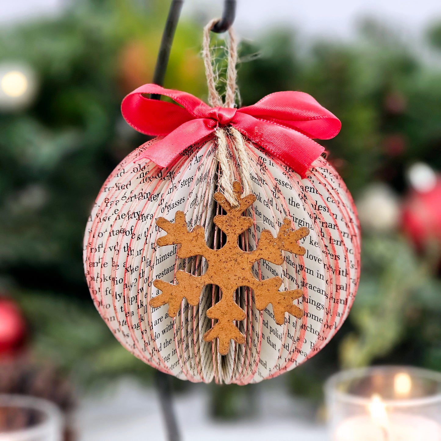 Round ball book page ornament made from book pages. It has a jute string for hanging, a red bow on the top and a rustic metal snowflake on the front.
