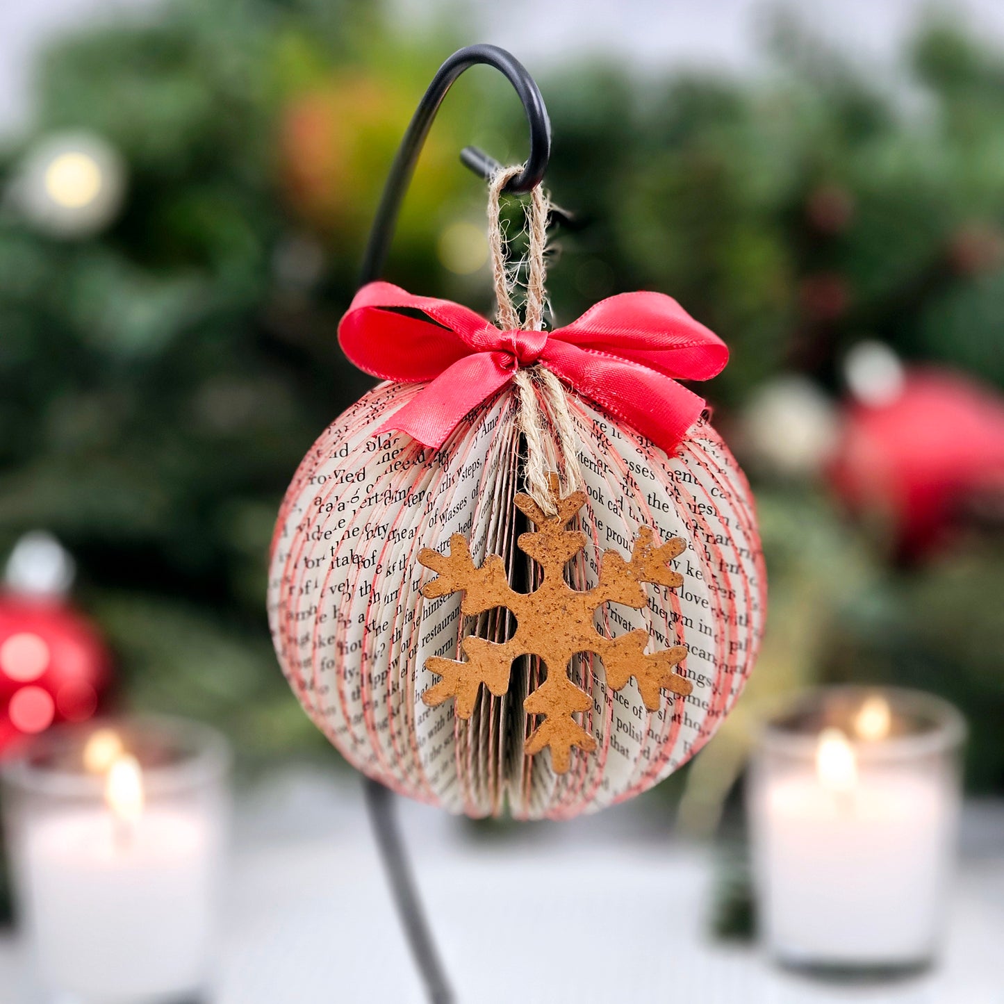 Round ball book page ornament made from book pages. It has a jute string for hanging, a red bow on the top and a rustic metal snowflake on the front.