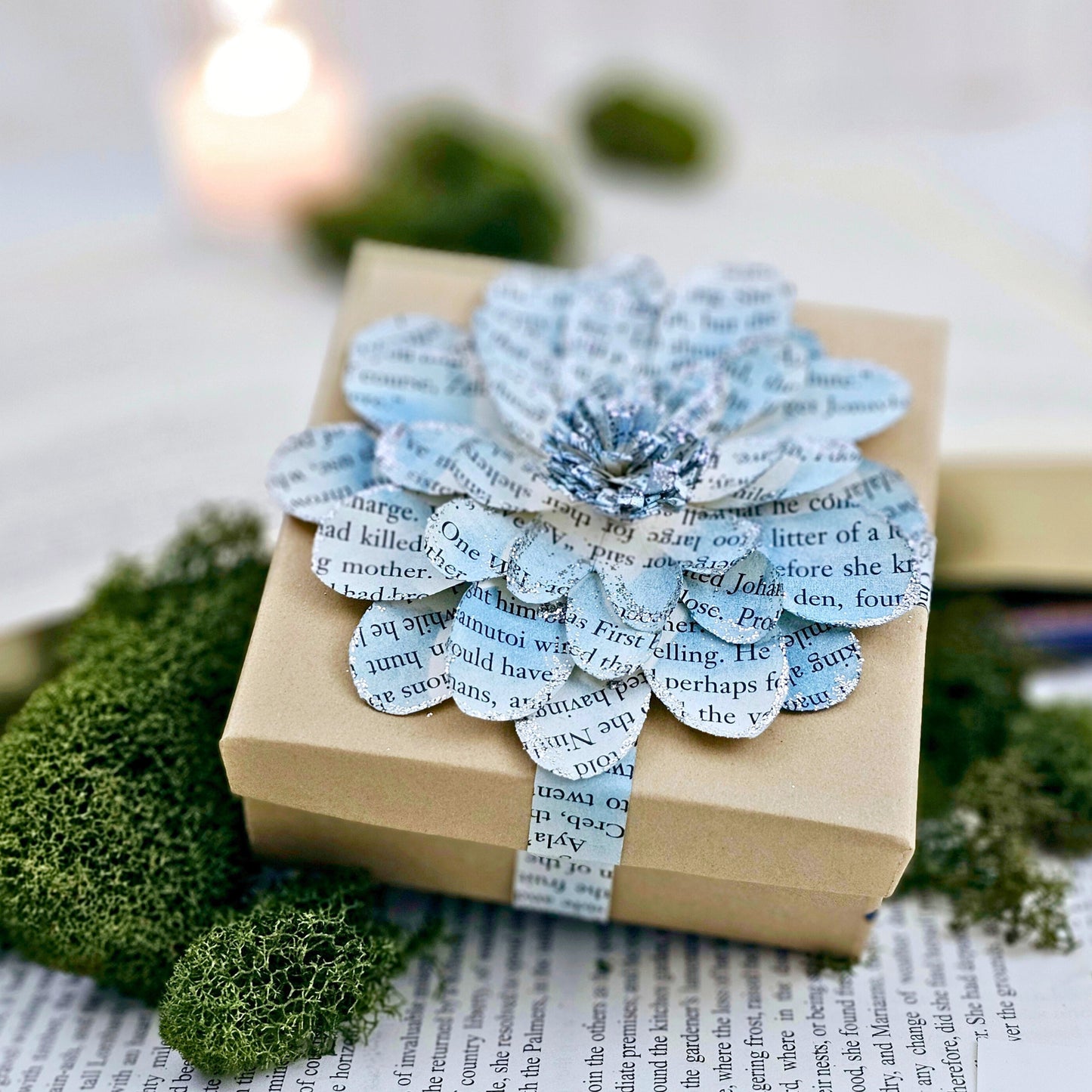 3.5 x 3.5 inch jewelry gift box adorned with a flower made from book pages. It is stained blue and has silver sparkly on the edges.