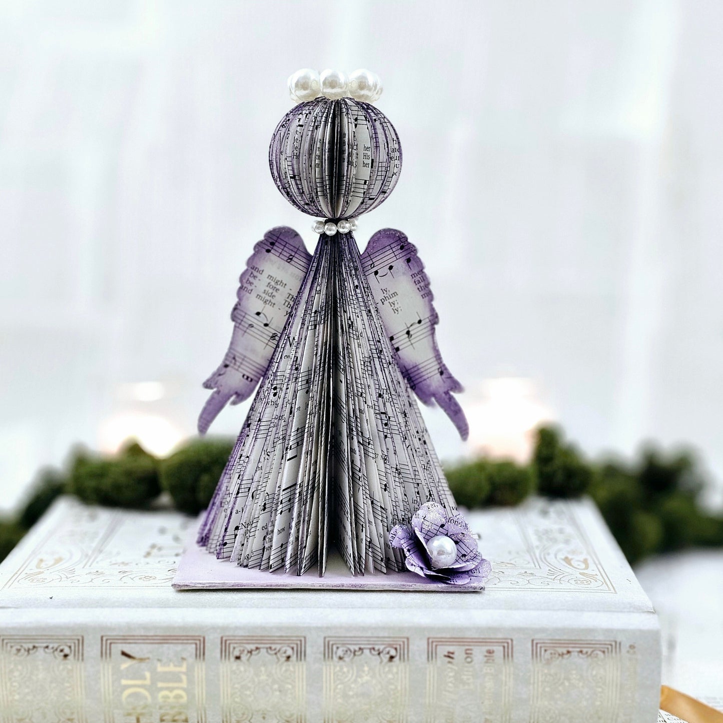 Angel made from hymn pages. The edges of the pages are stained purple. The wings also are stained purple on the edge and have a touch of sparkle on them. The angel wears a pearl halo and a dainty pearl necklace. The angel sits on a platform base made from the hymnal cover. The base has a flower on it made from hymn pages.