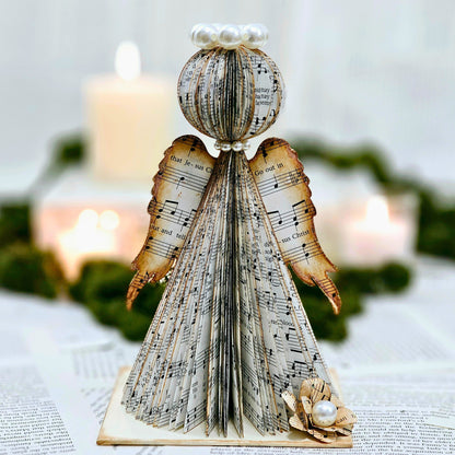 Angel made from hymn pages.  The edges of the pages are stained brown for a vintage aestethic.  The Edges of the wings have a touch of sparkle on them.  The angel has a crown of pearls and a dainty necklace of pearls.  The angel sits on a base made from the hymnal covers.  IT is painted beige and has a flower on it made from hymn pages.